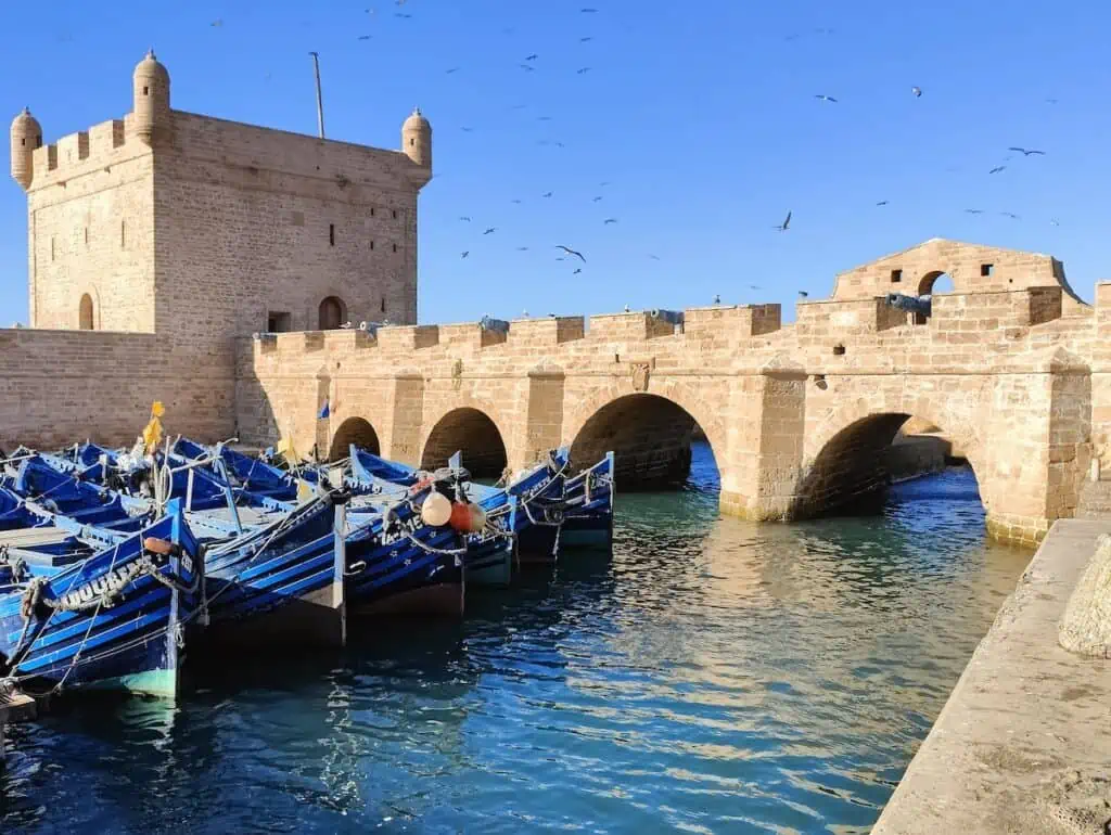 A view of blue boats sitting in the water on a sunny day. In the background, you can see an ancient stone, arches bridge leading to a square stone tower. This is what you'll see in the port during a Marrakech to Essaouira day trip.