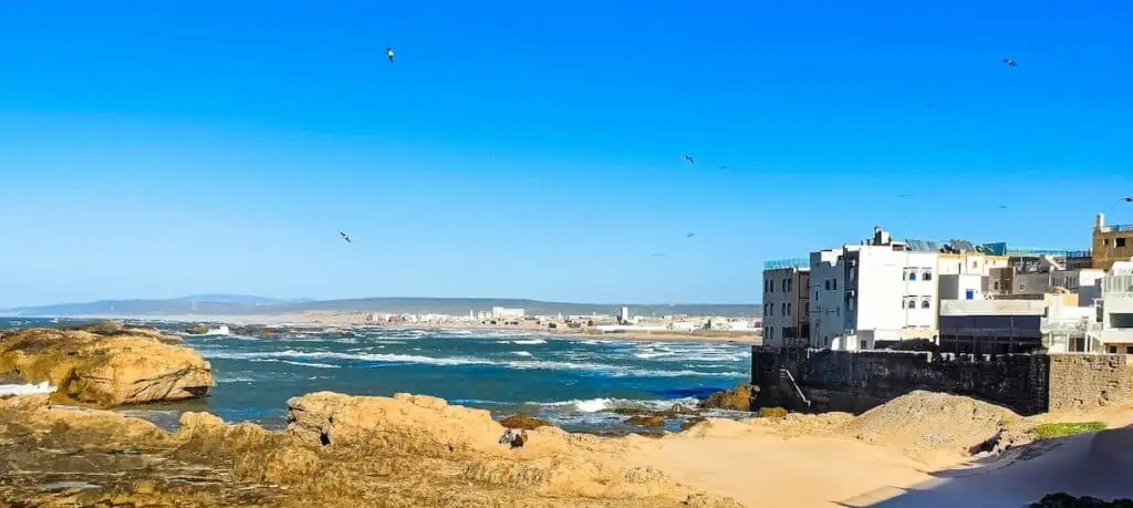 A view of the beach and the rocks that sit on the ocean in Essaouira. You can also see the white buildings and city on the edge of the sea.