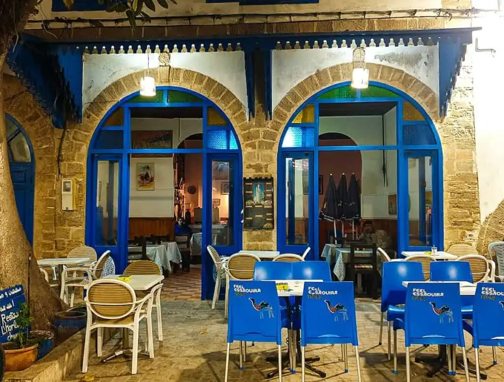 View of a blue and white restaurant with stone archways at night in Essaouira. Blue chairs and tables sit out front.