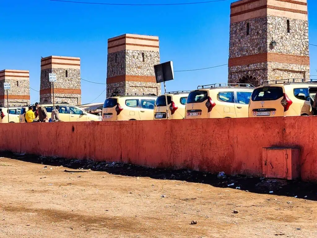 View of yellow grand taxis lined up against a wall with pillars behind them at the Grand Txxi station in Marrakech.
