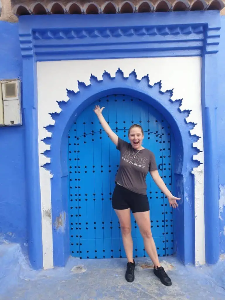 Me standing in front of a blue door in the Medina. I am wearing a gray t-shirt and black shorts and black sneakers. The door is round and has white trim. Consider the medina when deciding how many days in Chefchaouen.