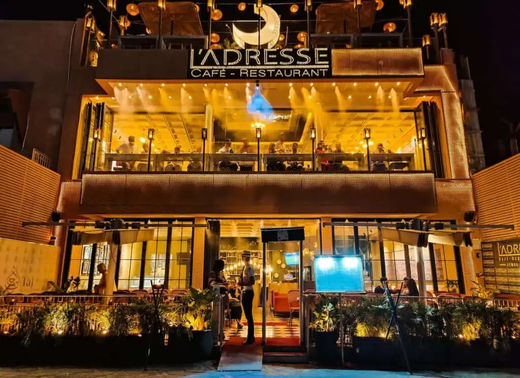 View of L'Adresse cafe all lit up in the evening with a sign on top of the two-storey restaurant with the name and moon lanterns hanging above, It sits in el Jemma fna square and is great place to enjoy one of the best things to do in Marrakech at night.