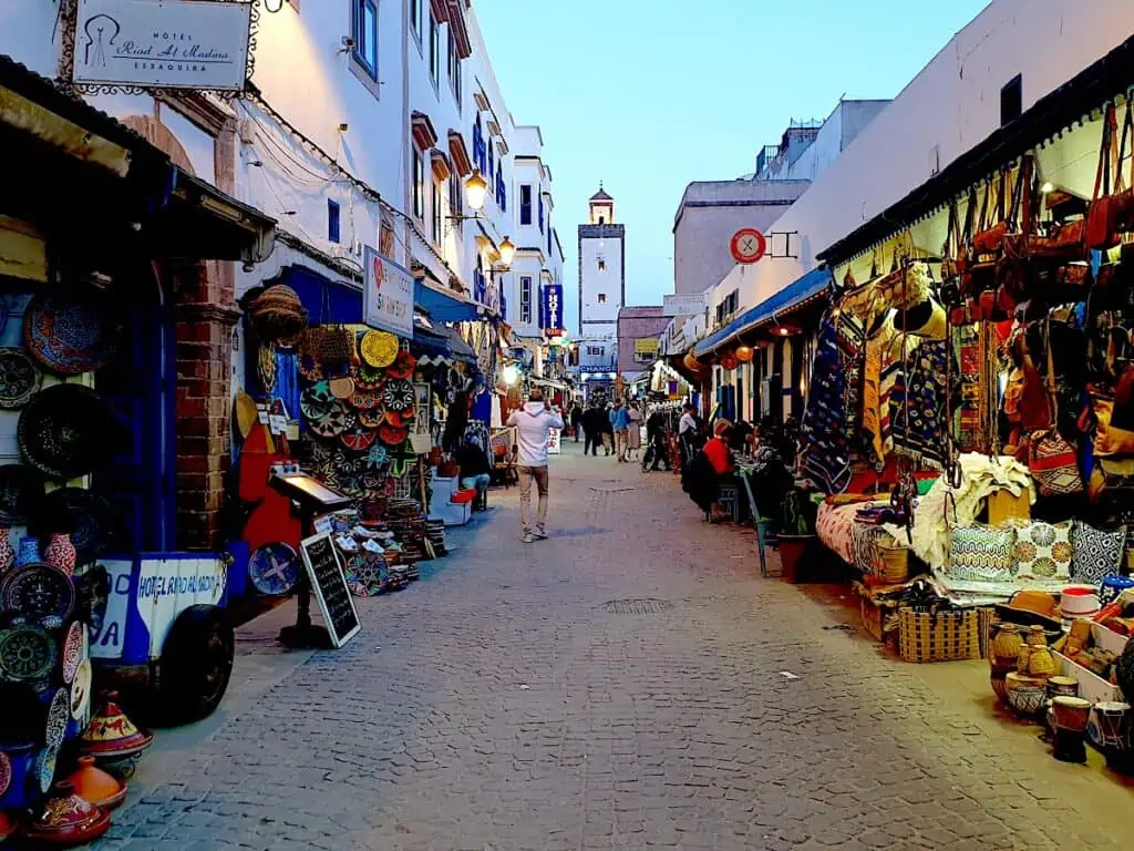 A view of the blue and white Medina with a white tower in the distance. People walk by local shops in Essaouira.