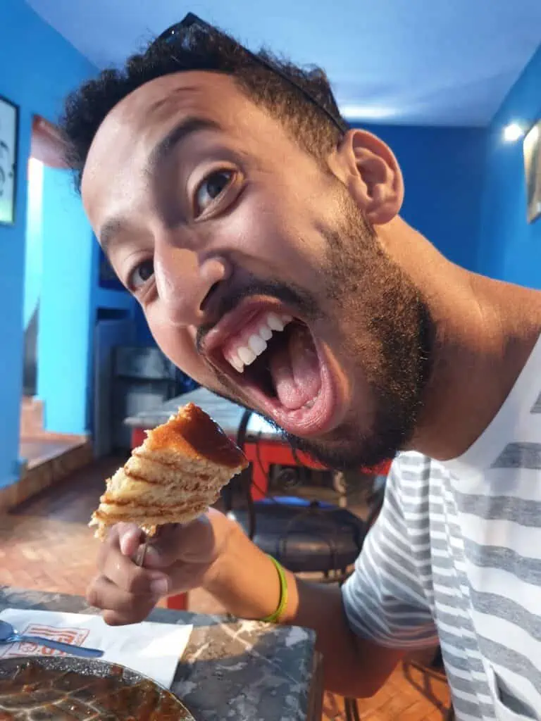 A close up of my boyfriend Rida eating pancakes on a fork with a blue wall in the background. He is wearing a white and gray striped shirt and has a fork with four triangular pieces of pancake on it.