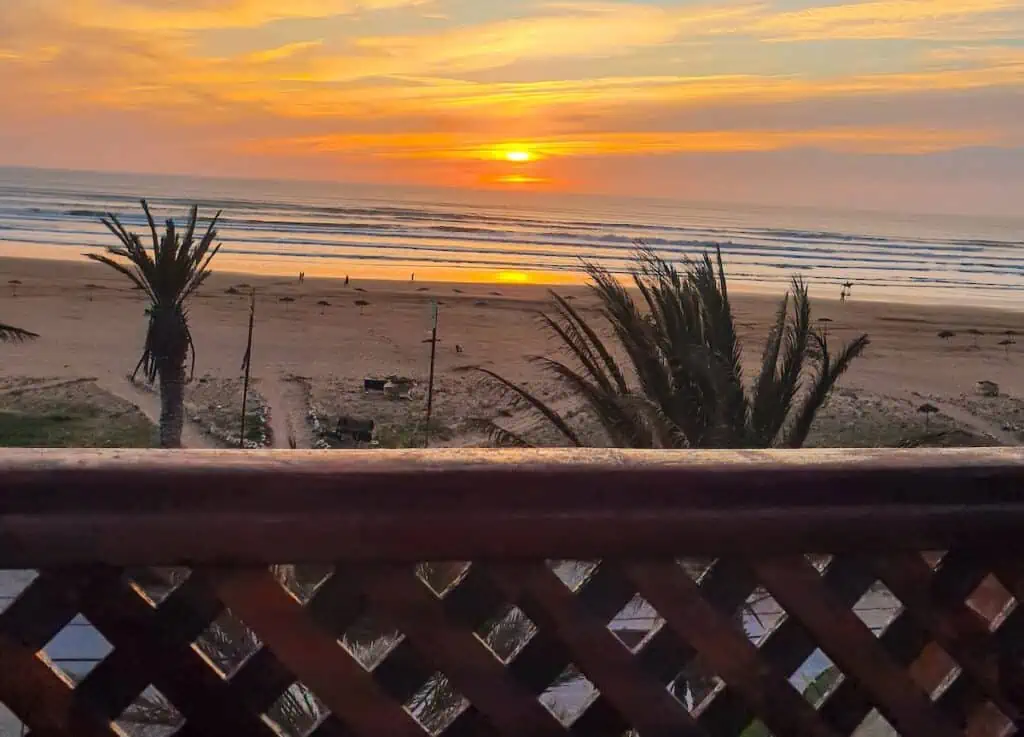 A view of the sunset in Sidi Kaouki while sitting behind a wooden railing with palm trees in front of the railing.