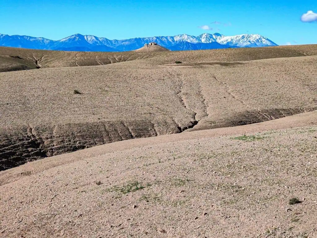 View of the rock hills in the Agafay Desert with a small community on a hill and the Atlas Mountains in the background on a sunny day.