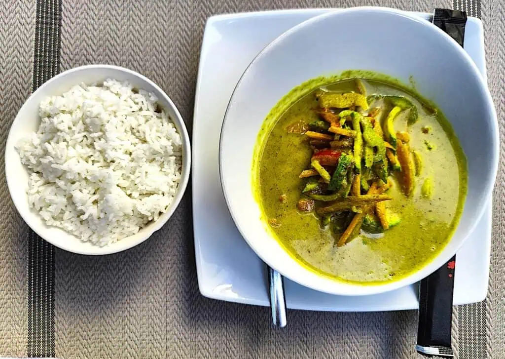 A view of vegetable green curry in a white bowl with a bowl of rice on the side on a gray placemat.