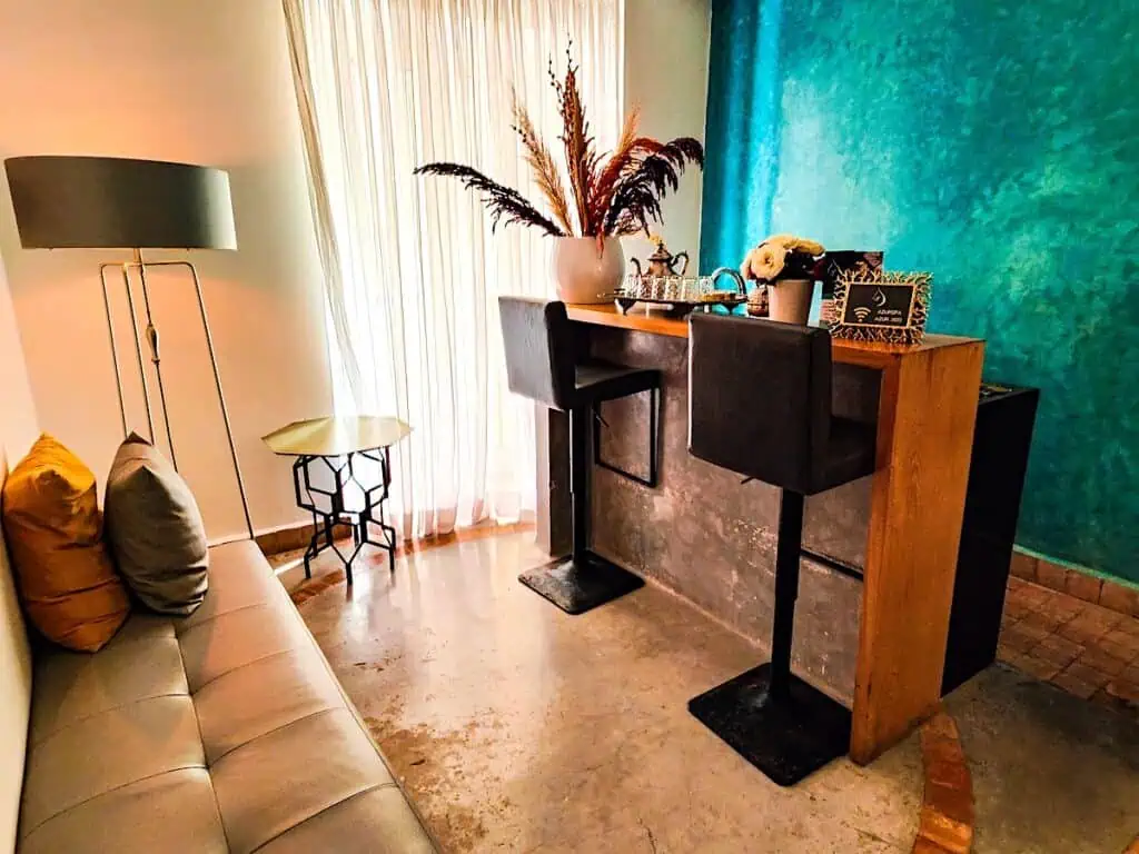 Room with a beige couch and a desk with plants on it. There is also a vibrant green wall in the back.