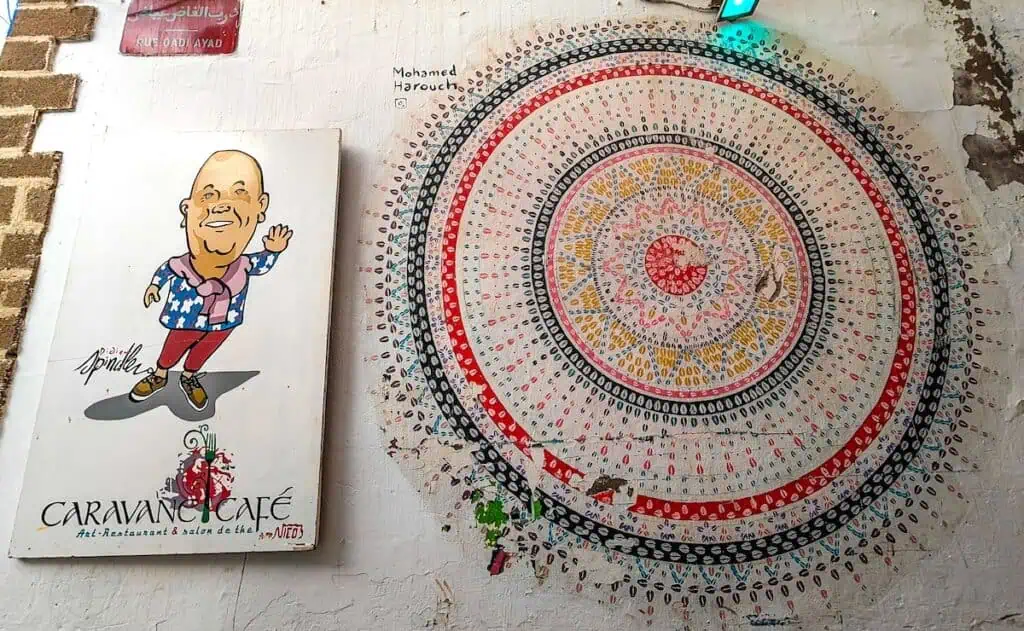 Cartoon of a man with w big, fat head on the left and a giant mandala on the wall on the right in Essaouira. It is a sign for Caravan Cafe in Essaouira.