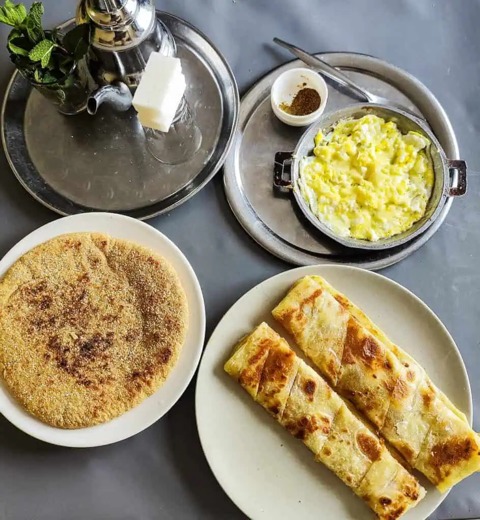Aerial view of a pot of tea, two msemen, bread, and an omelet iwht cumin on the side at a table in a breakfast place in Essaouira.