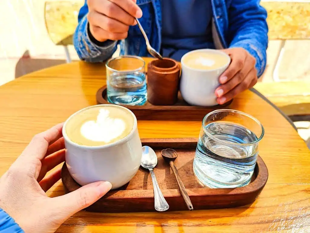 A tray with a latte, sugar, and glass of water. I am in the front and my partner is in the back.