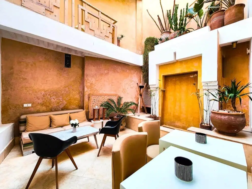 View of the common room with modern white tables and dark chairs, The roof is open and there are clay walls.
