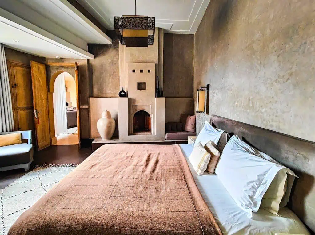 View of a large, full bed with a beige blanket and a fireplace in a gray wall in the background.
