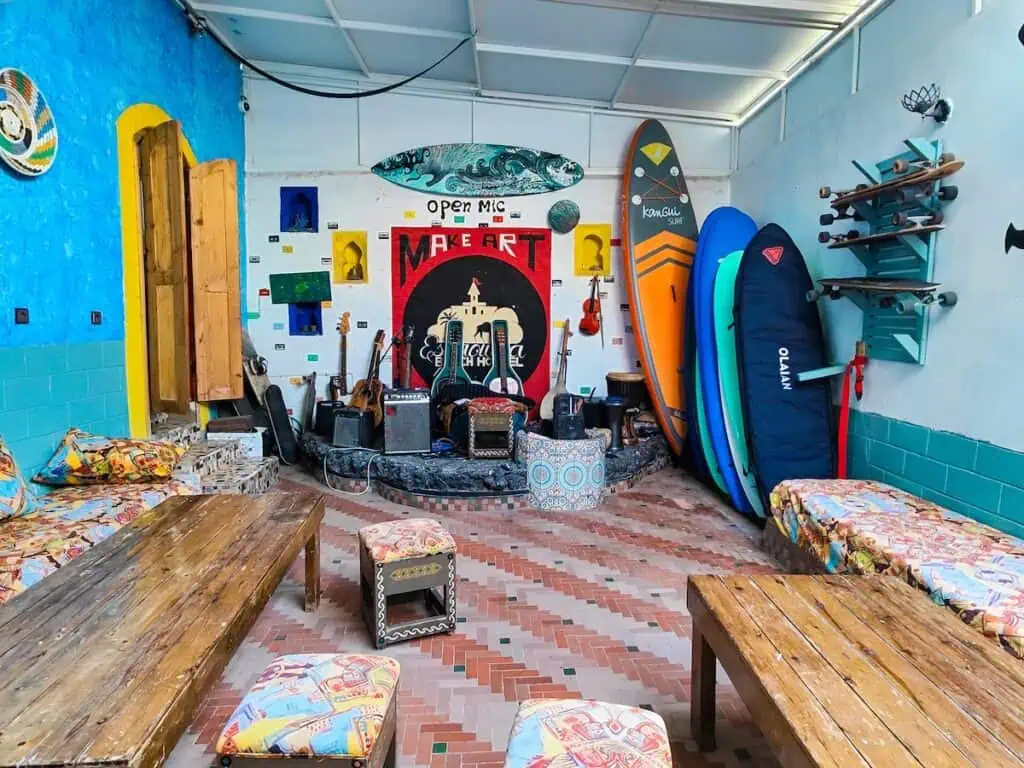 A common room with vibrant murals and a stage with guitars. Surboards also line the walls and you have long wooden tables and cushioned chairs where people can sit at one of the best hostels in Essaouira.