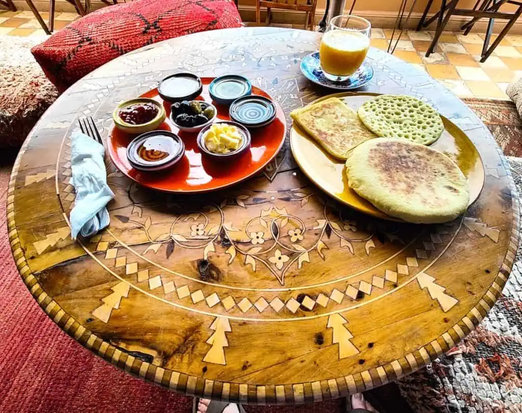 A plate of tradtitional Moroccan breads and a red plate filled with sides you can put on the bread. You also see cutlery in a napkin and OJ on a round, wooden table on the roof of KSOU.