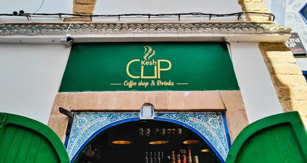 Green "Kech Cup" sign with gold letters above a blue doors with green shuetters and a coffee shop inside.