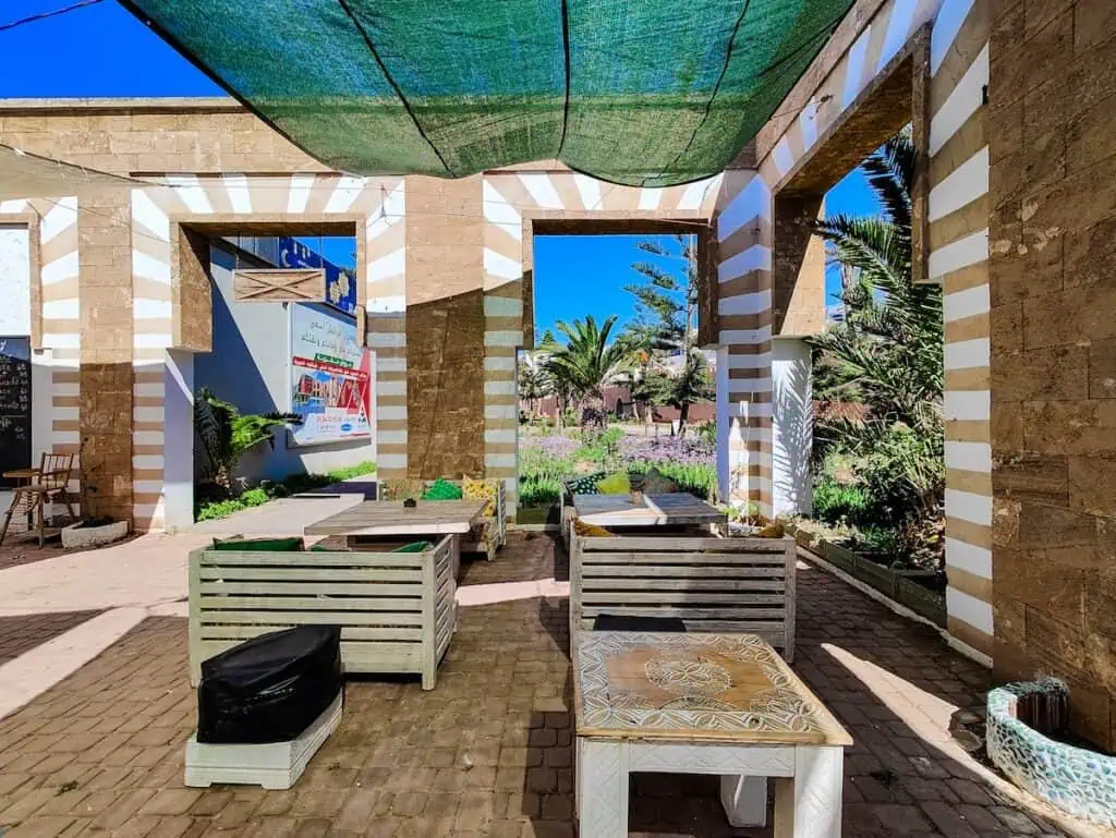 A view of the old brick building with disrressed white tables and chairs and large square open windows in the walls with green cloth hanging from the ceiling that makes up one of the best cafes in Essaouira.