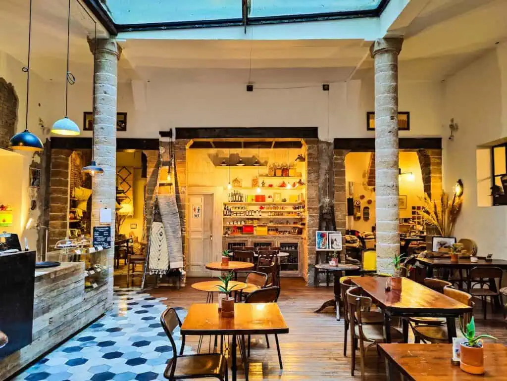 View of the light and airy dining room of L'Atelier with a sky light and tiled floors and wooden tables spread tables spread throughout the space.