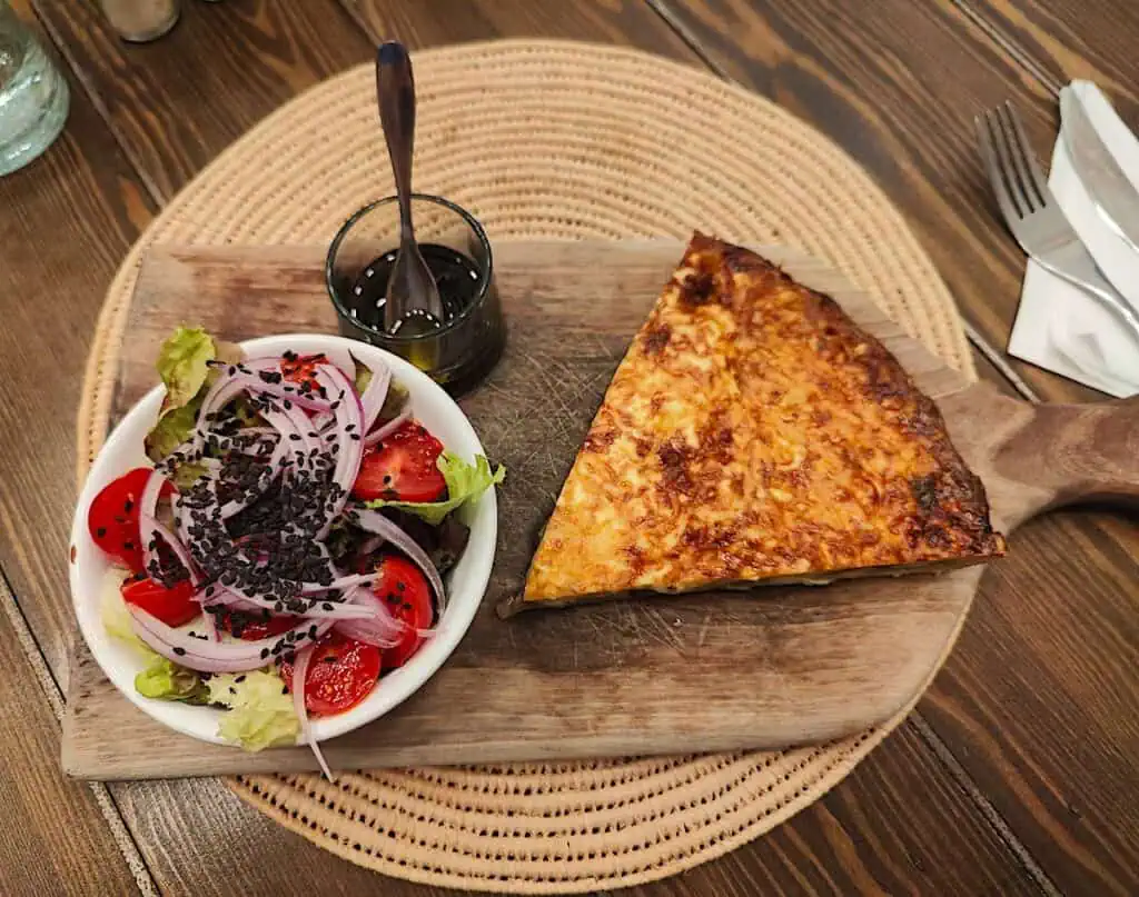 An aerial view of a slice of quiche and a salad with a side of vinaigrette on a wooden table.