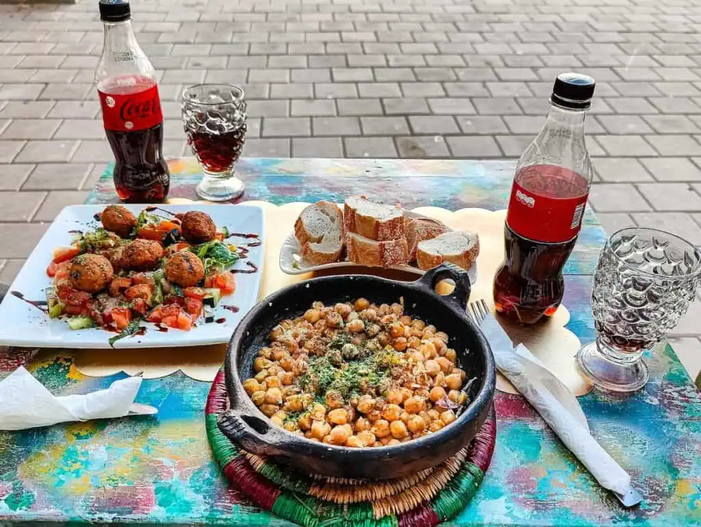 View of a multi-color table with a bowl of chickpeas and a plate of falafel with two bottles of Coke Zero and two glasses on the table.