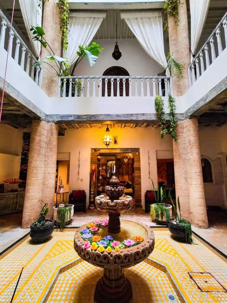 A view of the inner courtyard of the riad. You see w water fountain with flowers and rubber duckies with a yellow tile mosaic around it. There is a white wall on top and a railing with plants hanging down.