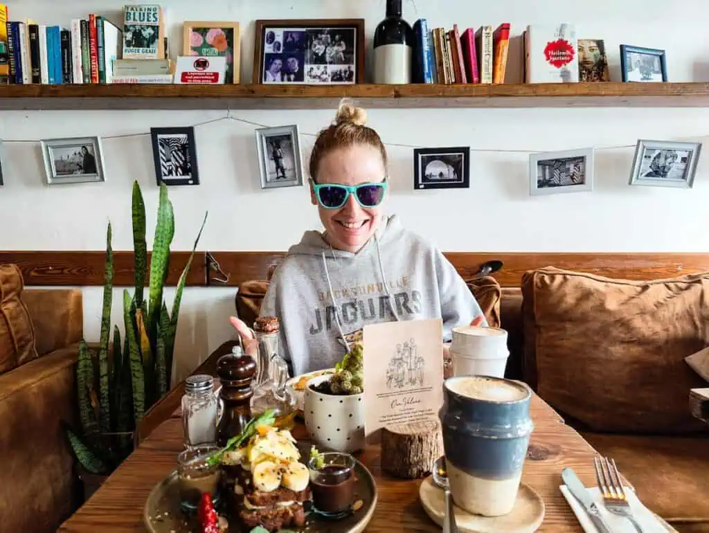 Me sitting in a gray sweatshirt and turquoise sunglasses on a brunch couch with hanging photos and a shelf of books behind me. You can see banana bread and avocado toast on the table as I enjoy the best breakfast Essaouira has to offer.