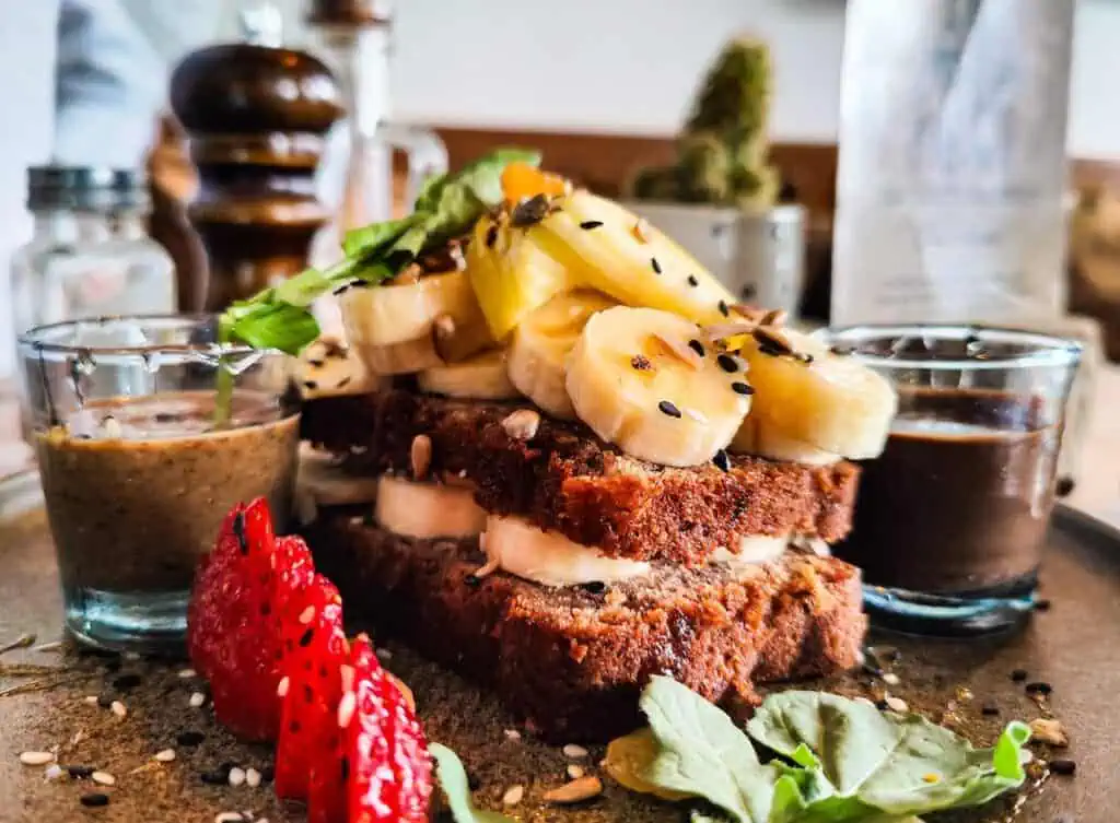 Up close view of banana bread with bananas on top and small cups of chocolate sauce and amlou on either side with strawberries in the front.