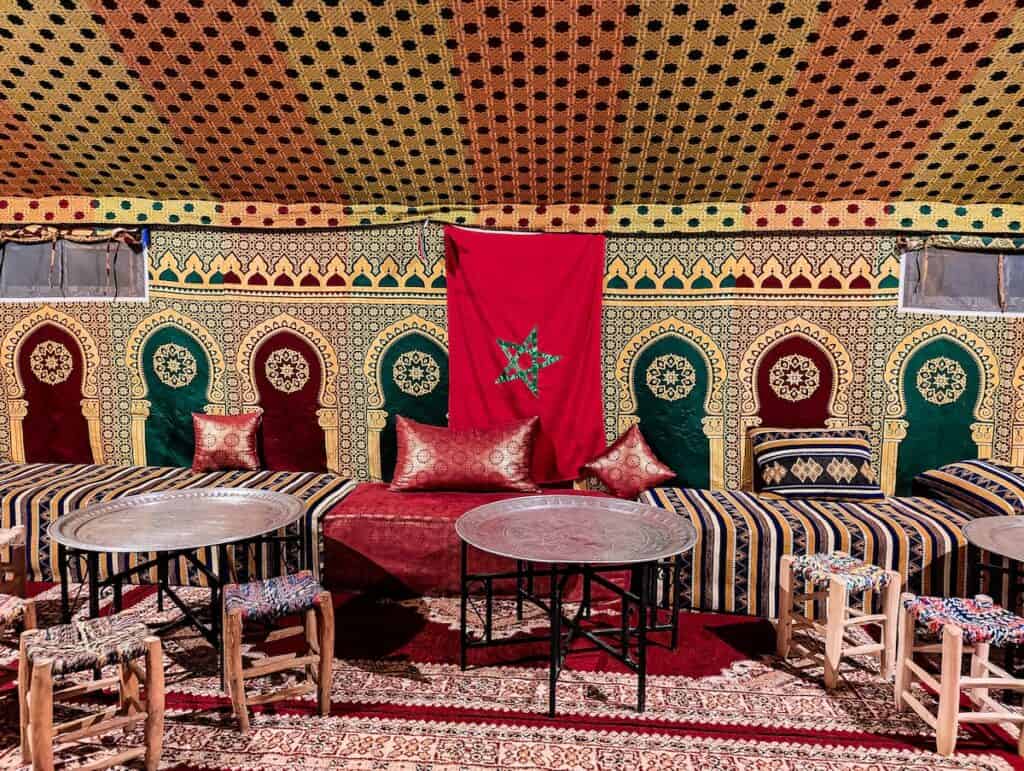 View of a Moroccan style tent with metal tables and vibrant seats with the red and green star of the Moroccan flag hanging in the background.