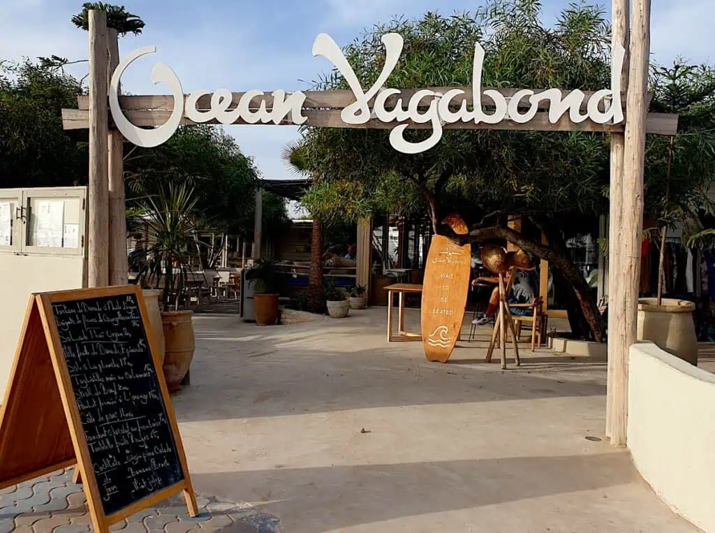 A view of the entrance sign that leads you into Ocean Vagabond. You can see a surfboard and tables and chairs and a menu on the left with a sign with white letters overhead that reads, "Ocean Vagabond".