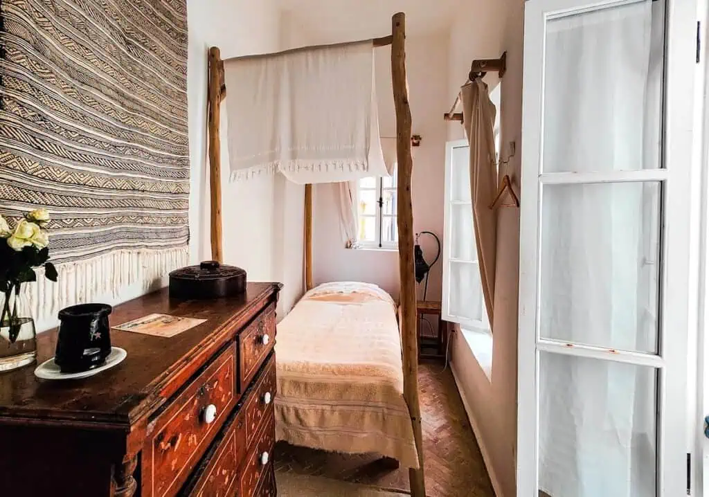 You can see a single bed with white sheets and a wooden dressed with wooden posts around the bed and a beige rug hanging on the wall/