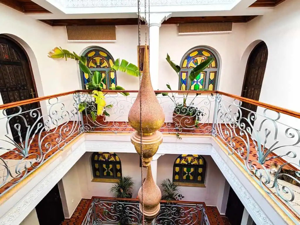 View of a open courtyard in a riad with white walls, wooden banisters, and clay lamps hanging from the ceiling. You can see multi-color arched windows in the background.