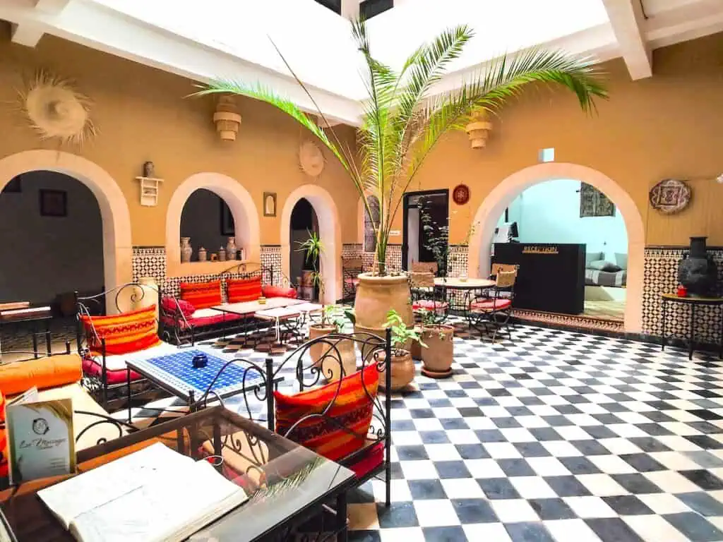 Vast courtyard inside a riad with bleck and white tile floors, large potted plants and red chairs around tables.