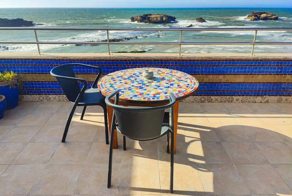 View of the beach and an island in the distnace from the roofop terrace of Riad Perle D'eau. You can see a round table with a multi-color tabletop in the sun looking out at the ocean with two chairs around it.