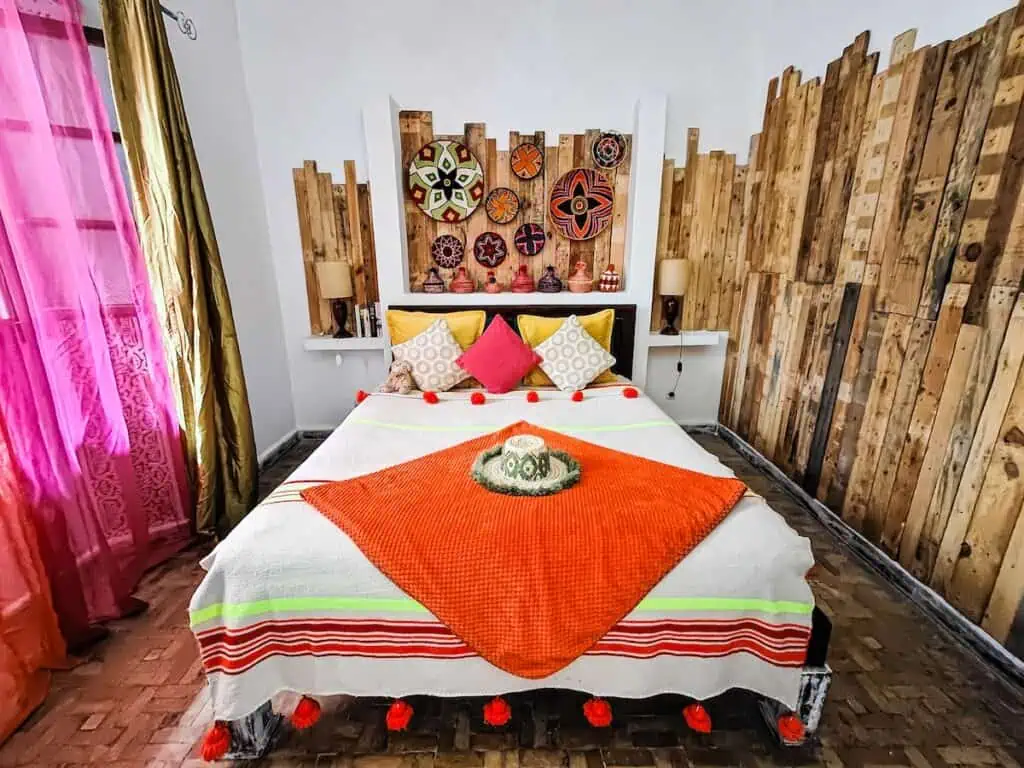 View of a full size bed with a white bed spread and an orange blanket. You can see wood pannels in the background with circuliar vibrant decorations on the wood board in the back. There is wood on one side and a pink curtain on the other at one of the best luxury riads in Essaouira.