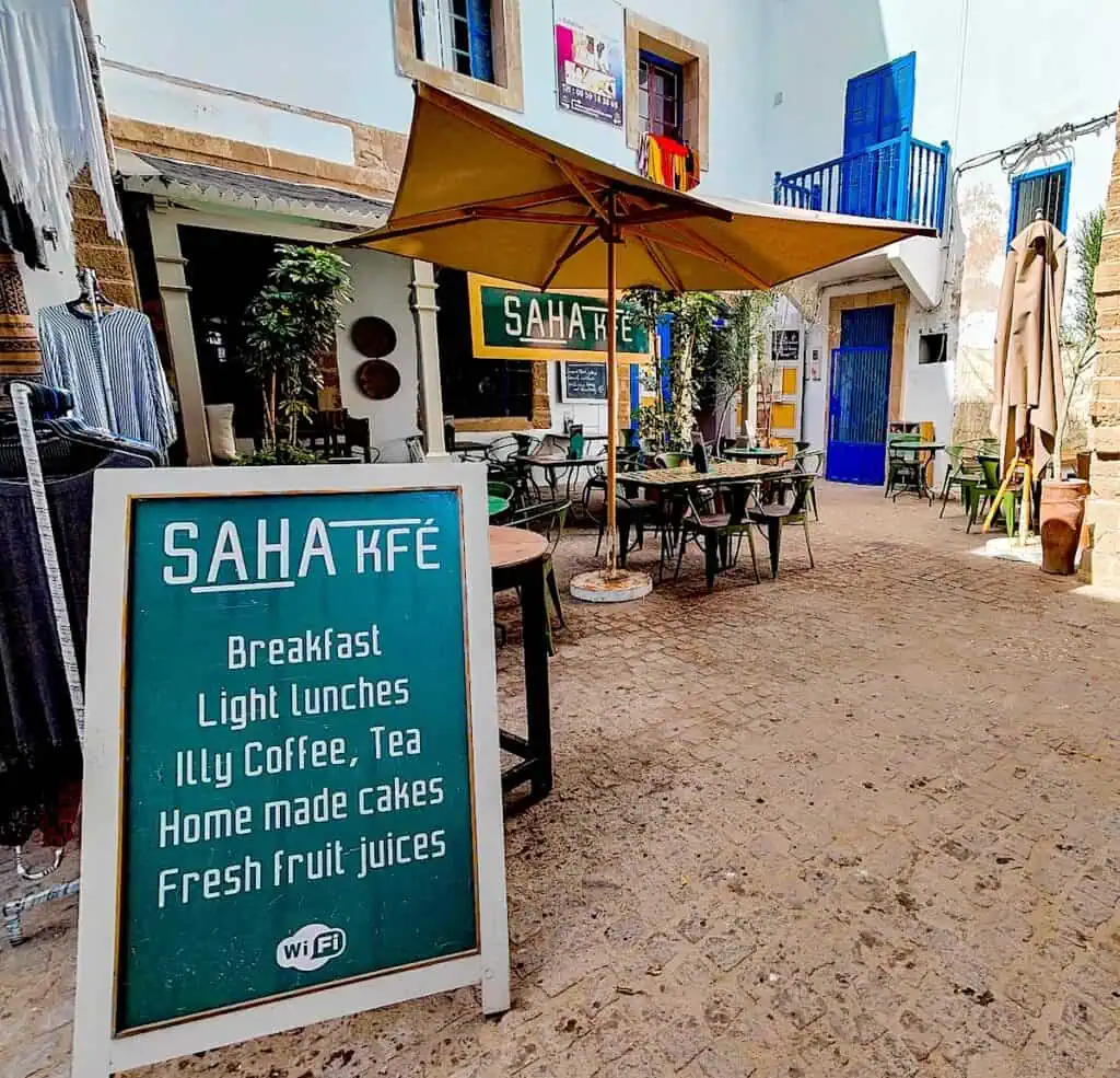A view of the outdoor in Essaouira with tables and chairs and a nice umbrella above it. There is alsoa. green standing sign with the name of the cafe on it.