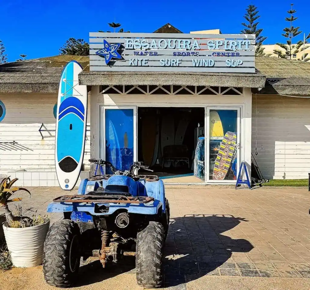 Entrance into Essaouira Spirit surf shop along the beach promenade in Essaouira. You see a blue surf board standing on the left side of the entrance and a blue quad bike sitting out in front off the entrance of the white building.