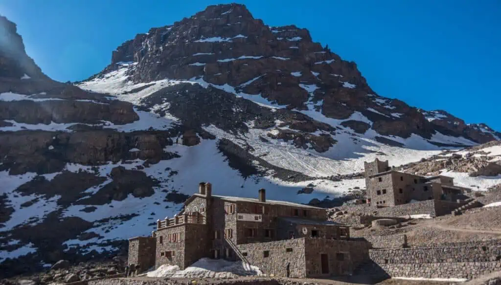 View of Mount Toubkal covered in snow with a stone house at the base of the mountain.