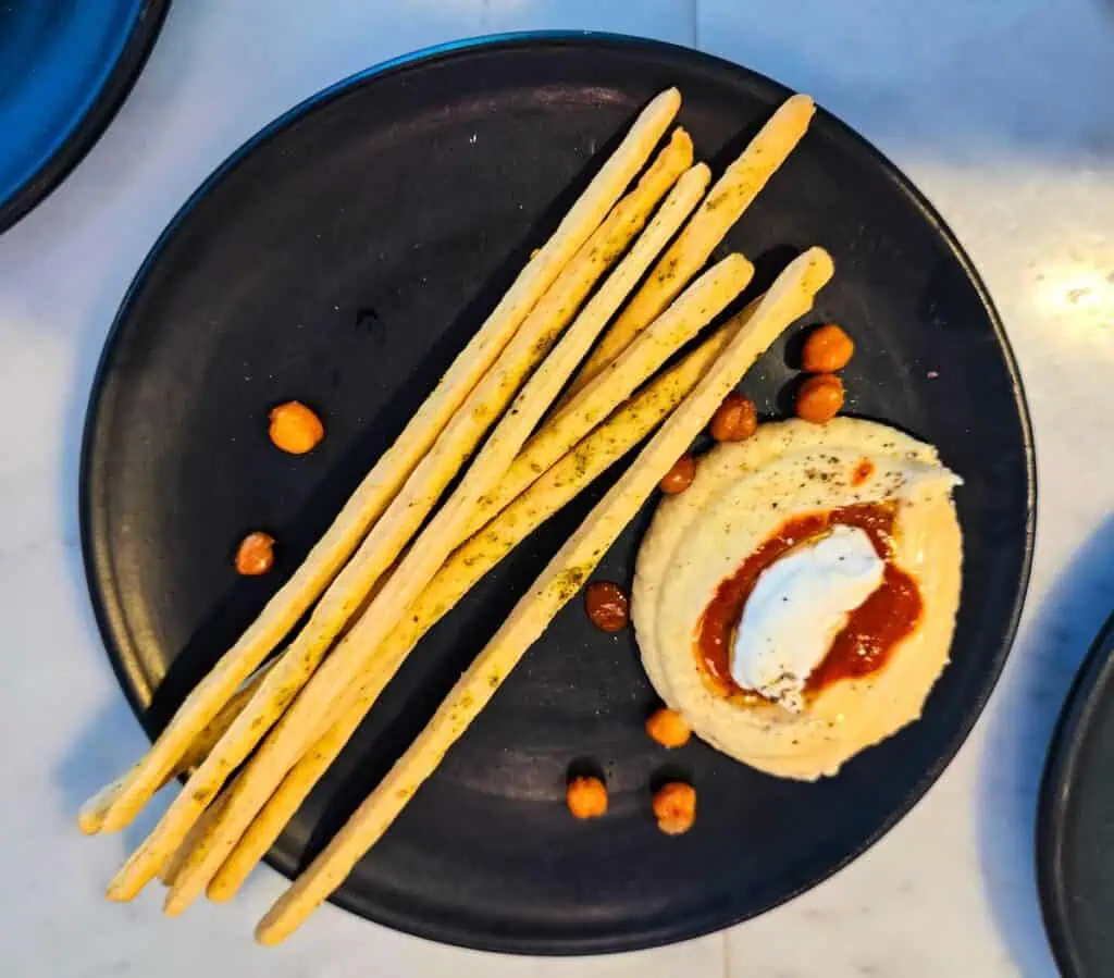 Aerial view of a black plate with thin breadsticks going across the plate. You have a mound of beige hummus on the right with white cheese and red sauce on top at one of the best restaurants in marrakech.