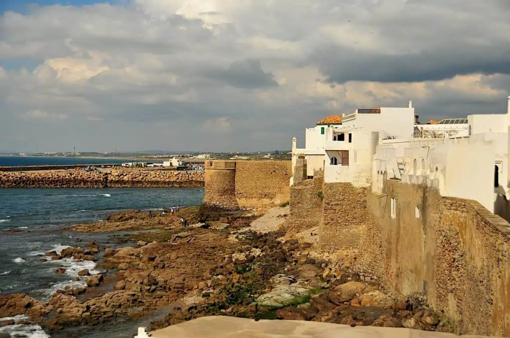 View of the white buildings and steep stone walls that sit along the coast of the ocean in Asilah in Morocco. it's one of the prettiest cities in Morocco.