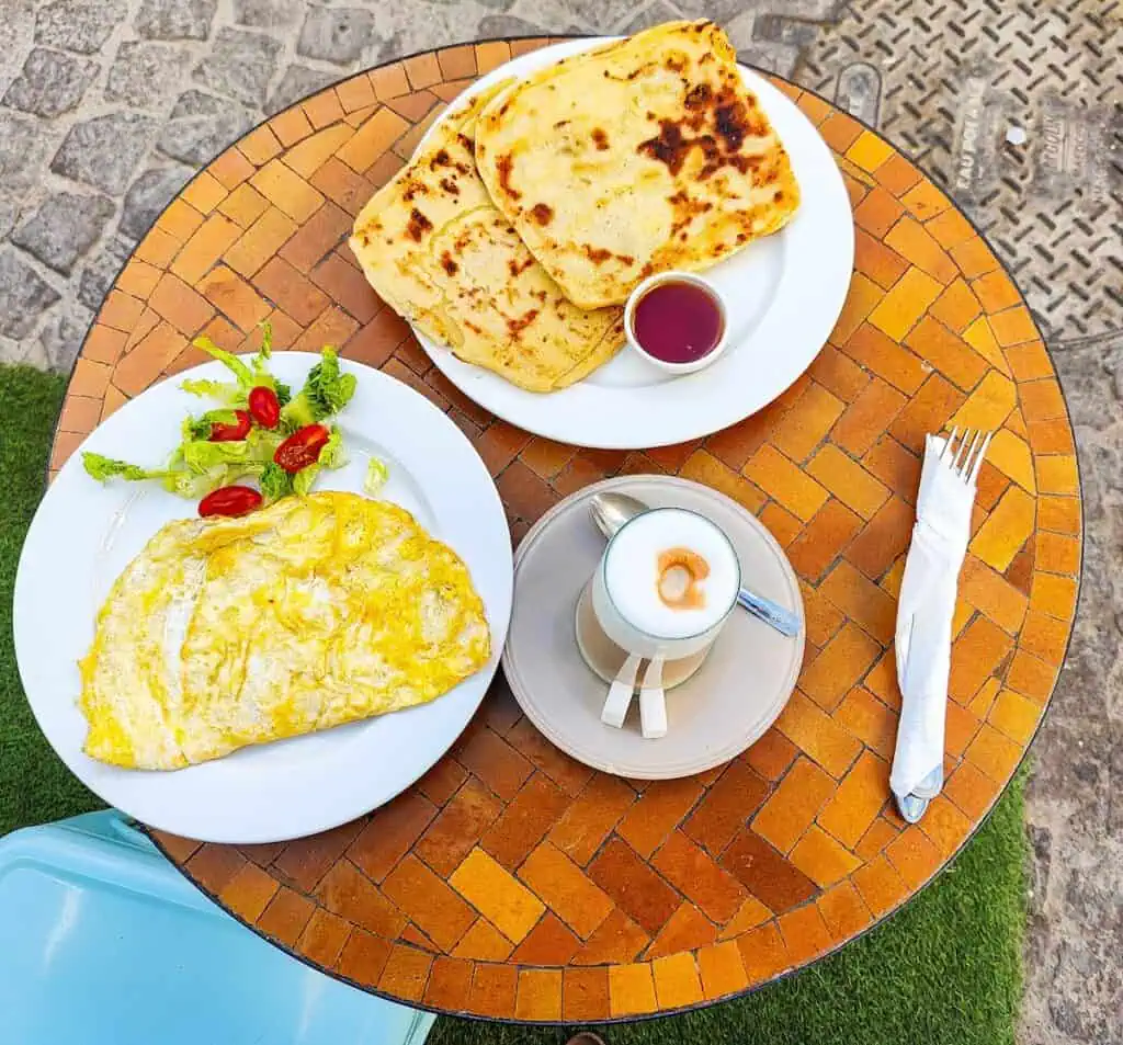 Aerial view of a brown, tile table with a set of silverware in a white napkin on the right, a latte with white foam on a gray plate with a spoon in the middle, an omelet on a white plate with a side salad on the left, and two msemen with a side of honey on a white plate at the front of the table.