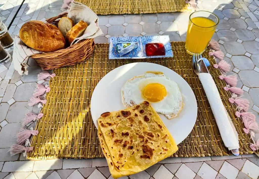 View of breakfast at Sun Hostel. It includes an egg, msemen, a croissant, bread, cheese, jam, and juice on a wooden place mat.