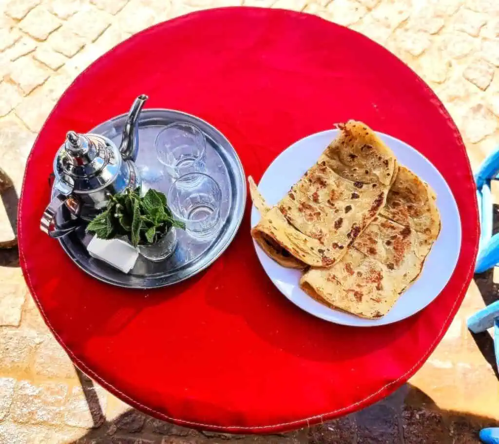 Aerial view of a red, round table. On the right you have two msemen folded in half on a white plate and on the left you hav e a pot of two, two glasses, sugar, and cup of mint on a silver tray.