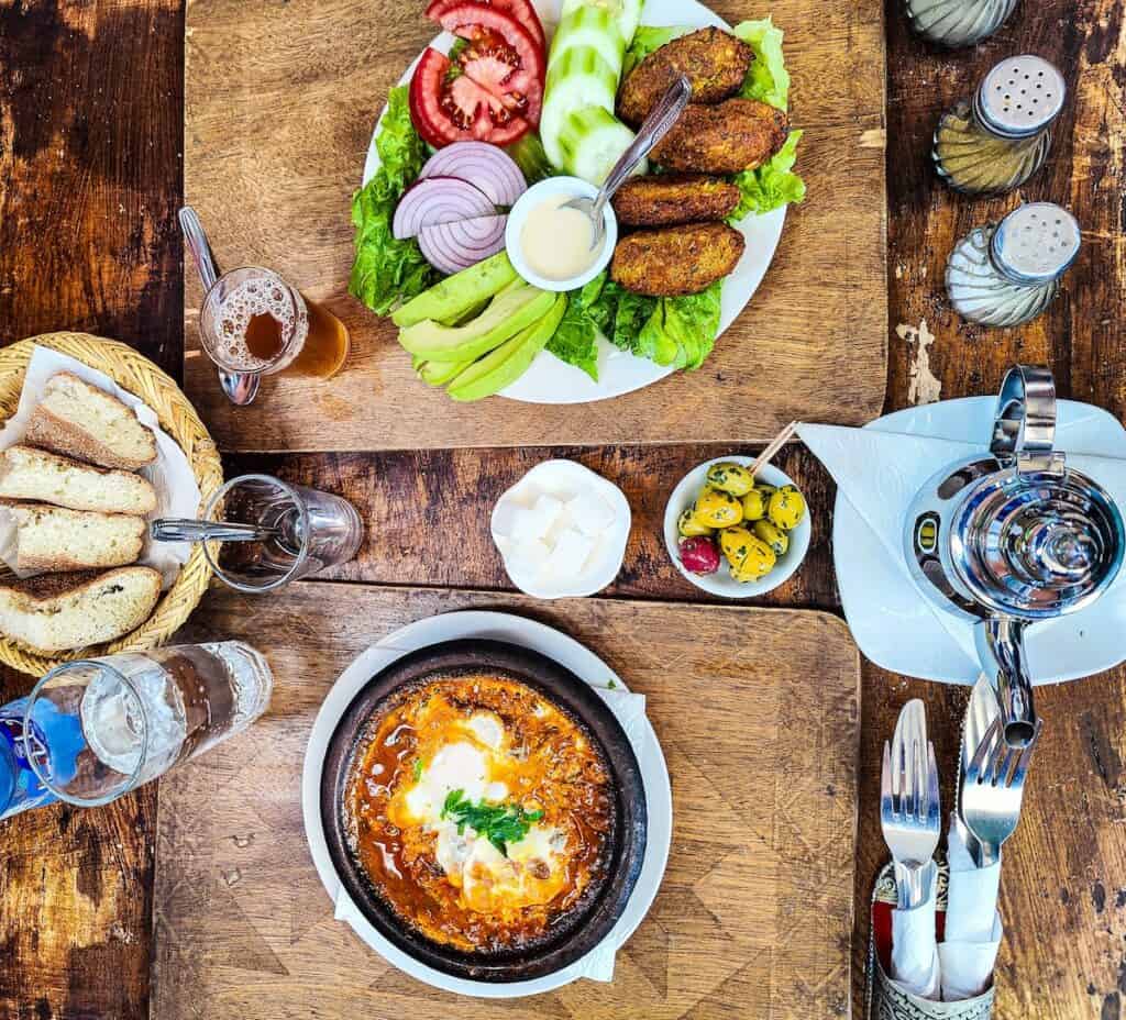 Aerial view of food on a wooden table. You can see a Berber Omelet, a falafel plate, a bowl of olives, a pot of tea, and a basket of bread at Henna Art Cafe.