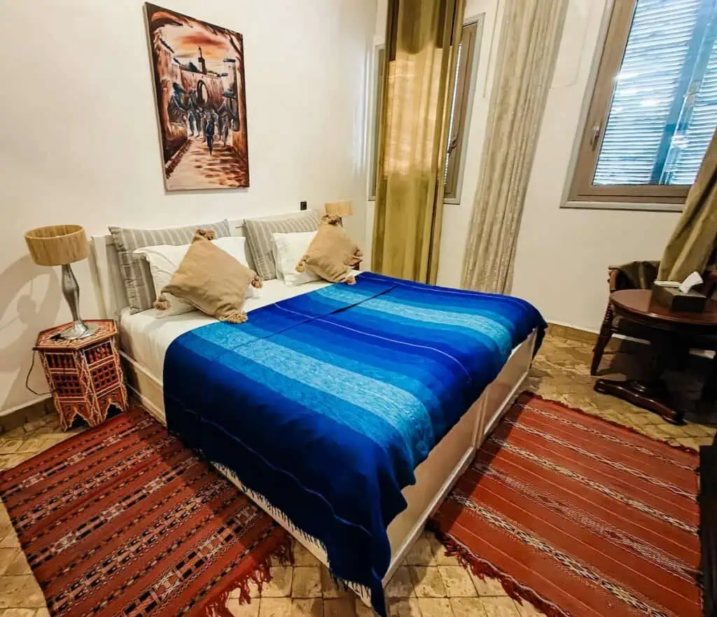 View of a tradtional bedroom inside of Koutoubia Royal Riad. You can see Berber rugs on the floors and a bed with a blue blanket and white walls.