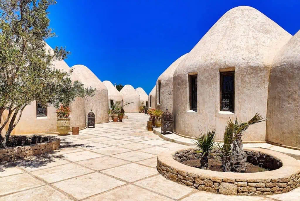 View of the white huts with a triangular roof and inset windows at Les Jardins de Villa Maroc on a sunny day with a tiled walkway.