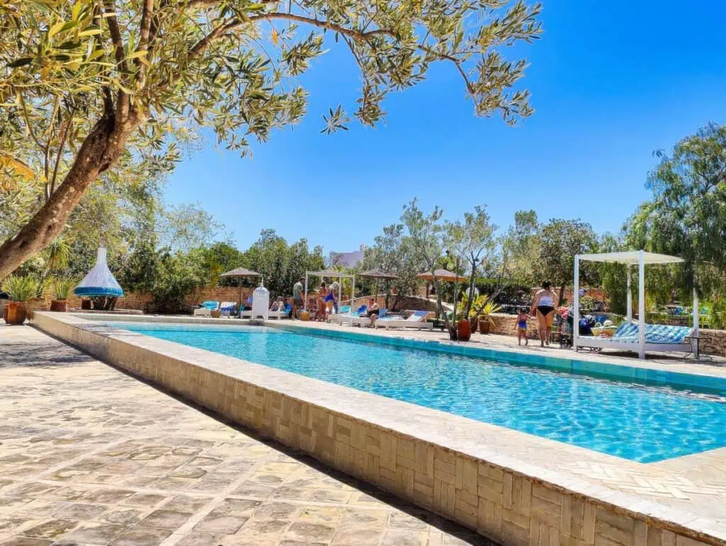 A view of the large, rectangular pool at Les Jardins Villa de Maroc. A tree is on the top left and it is a sunny day. You can see cabanas around the pool.