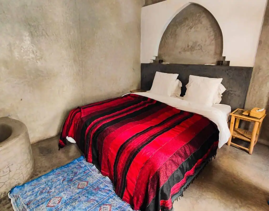 View of a big bed with a red blanket and a blue rug on the floor.