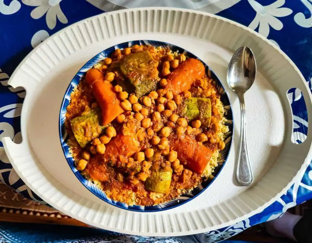 Aerial view of a plate of vegetarian couscous with chickpeas, carrots, and zucchini in a bowl with a spoon on the right. Its sits on a blue table cloth and part of a cooking class that is one of the best things to do in Essaouira at night.