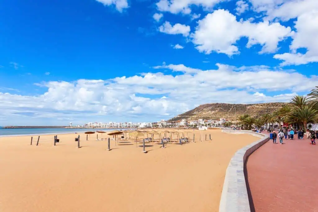 A view of the main beach in Agadir with yellow sand, hills in the background, and a brick promenade to the right.
