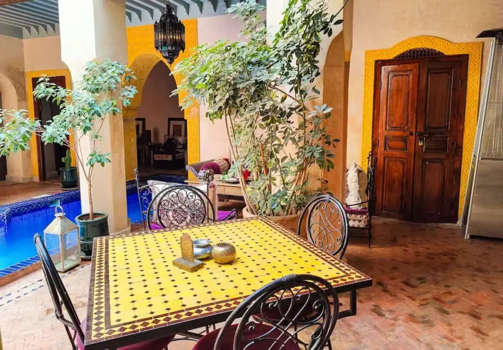 The inner courtyard of Riad Bayti with yellow tile tables and a blue pool as well as potted plants and yellow accents around the doors.